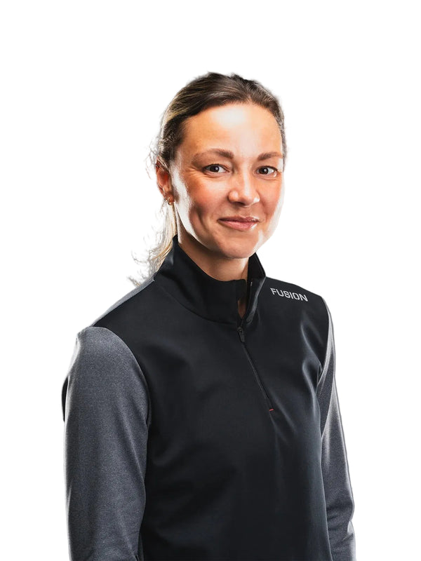 En kvinde med brunt hår, der er sat op, iført Fusion Tempo! Shield Zip til kvinder i mørkegrå, står med ansigtet vendt fremad foran en hvid baggrund.