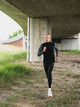 En mand iført en mørkegrå Fusion Tempo! Shield Zip, Herre og sorte sportsleggings løber på en grussti under en betonbro.