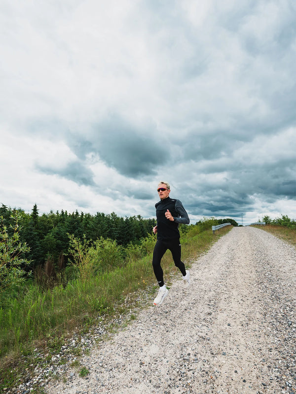 En mand iført en sort Fusion Tempo! Shield Zip, Herre, mørkegrå og leggings løber ad en grusvej under en overskyet himmel.