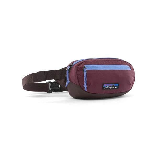 En Patagonia Terravia Mini Hip Pack i farvekombinationen Berry Fig har en blå lynlås, en justerbar rem og det karakteristiske Patagonia-logo på forsiden. Denne unisex-bæltetaske kan også bæres som en skuldertaske og er her afbilledet i en let vinkel på en hvid baggrund.