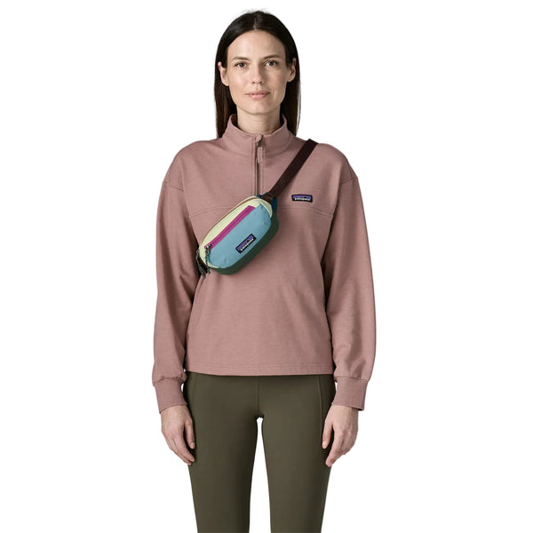 En person står med ansigtet vendt fremad og er iført en lilla sweatshirt med kvartlængde lynlås, olivengrønne bukser og en lille Patagonia Terravia Mini Hip Pack (unisex, patchwork: Snowmelt Blue), som bæres på tværs af kroppen. Baggrunden er ensfarvet hvid.