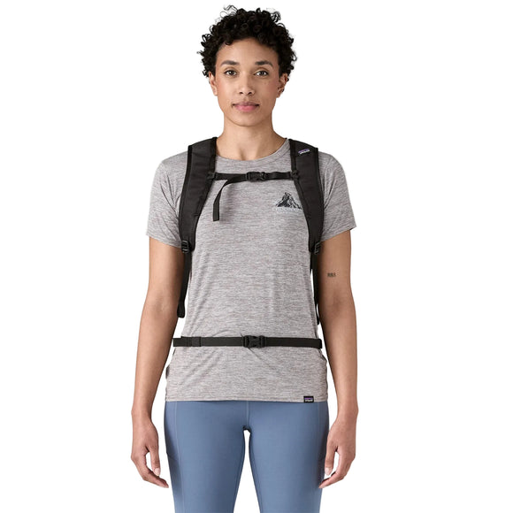 En person med kort, krøllet hår har en grå T-shirt, blå leggings og en sort Patagonia Terravia Pack 14L med bryst- og taljestropper på og står foran en ensfarvet hvid baggrund.