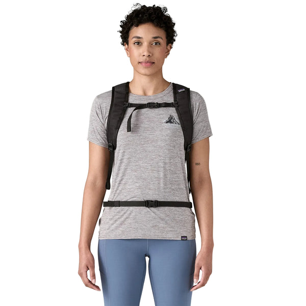 En person med kort, krøllet hår har en grå T-shirt, blå leggings og en sort Patagonia Terravia Pack 14L med bryst- og taljestropper på og står foran en ensfarvet hvid baggrund.