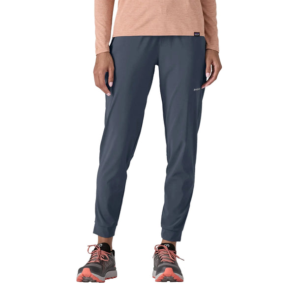 En person er iført en langærmet ferskenfarvet top, Patagonia Terrebonne Joggers til kvinder i Smolder Blue og grå trail-løbesko med orange detaljer. Billedet er beskåret fra skuldrene og ned på en hvid baggrund.