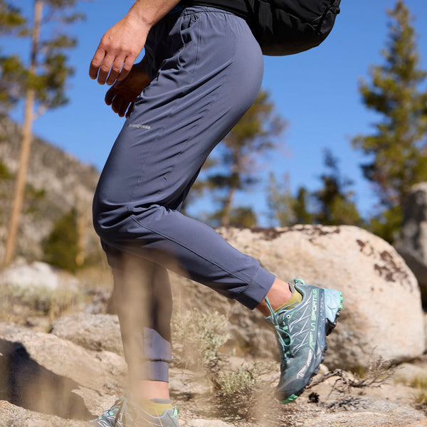 En person vandrer i stenet terræn blandt fyrretræer iført Patagonia Terrebonne Joggers, Dame i Smolder Blue, blågrønne trailsko og en sort rygsæk. Store klipper er synlige i baggrunden.