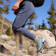En person vandrer i stenet terræn blandt fyrretræer iført Patagonia Terrebonne Joggers, Dame i Smolder Blue, blågrønne trailsko og en sort rygsæk. Store klipper er synlige i baggrunden.