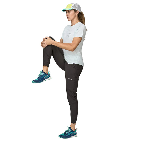 En kvinde i sportstøj og en kasket står på sit højre ben, mens hun holder sit venstre knæ op mod brystet, som om hun strækker ud. Hun har en lys trøje på, et par sorte Patagonia Terrebonne Joggers til kvinder og blågrønne løbesko. Hvid baggrund.