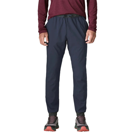 En person står med ansigtet vendt fremad og er iført en rødbrun langærmet skjorte, mørkeblå Patagonia Terrebonne Joggers, Herre (New Navy), fremstillet af genanvendt polyester, samt sorte og lyserøde trail-løbesko. Billedet er beskåret ved halsen, og personens ansigt ses ikke.