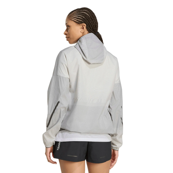 En person står med ryggen til og har en Terrex Xperior Light Windweave-jakke til kvinder fra Adidas på, i sort og hvidt, med lange ærmer, synlige sømme, mesh-detaljer og et elegant design. Personen har desuden sorte sportsshorts på foran en ensfarvet hvid baggrund.