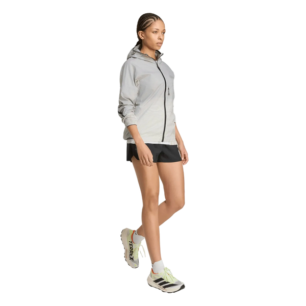 En kvinde iført en Adidas Terrex Xperior Light Windweave-jakke til kvinder i sort og hvidt, kombineret med sorte shorts og hvide løbesko med sorte og neongrønne detaljer, går fremad foran en ensfarvet hvid baggrund.