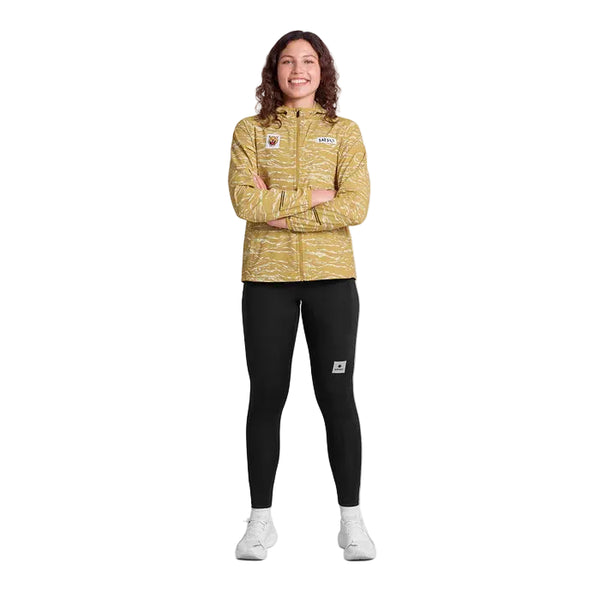 En person står med ansigtet fremad og armene over kors, iført Saysky Tiger Pace Jacket, Dame, Yellow Camo for optimal vindbeskyttelse, sammen med sorte leggings og hvide sneakers på en almindelig hvid baggrund.
