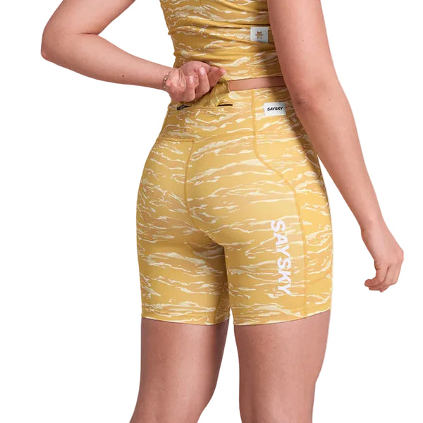 En kvinde iført Saysky Tiger Combat+ Short Tights 7" i Yellow Camo, fremstillet af genbrugspolyester, ses bagfra i færd med at justere sin matchende top. "SAVSIV" er trykt i hvidt på shortsene.