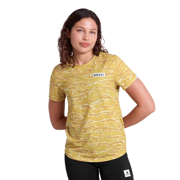 En kvinde med krøllet brunt hår bærer en Saysky Tiger Combat T-shirt, Dame, Yellow Camo, med et brystmærke, og sorte bukser, mens hun står med hænderne på ryggen mod en hvid baggrund.