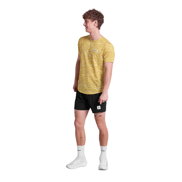 En mand står lidt til venstre og smiler i en Saysky Tiger Combat T-Shirt, Herre, Yellow Camo, sammen med sorte shorts, hvide sokker og hvide sneakers på en almindelig hvid baggrund.