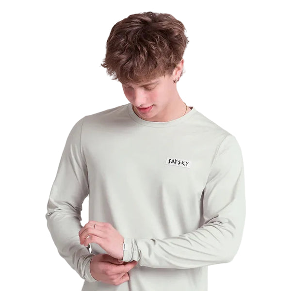 En ung mand med kort, bølget brunt hår bærer Saysky Tiger Pace Long Sleeve, Herre, Grey med SAYSKY på brystet. Han kigger ned og justerer sit ærme mod en hvid baggrund.