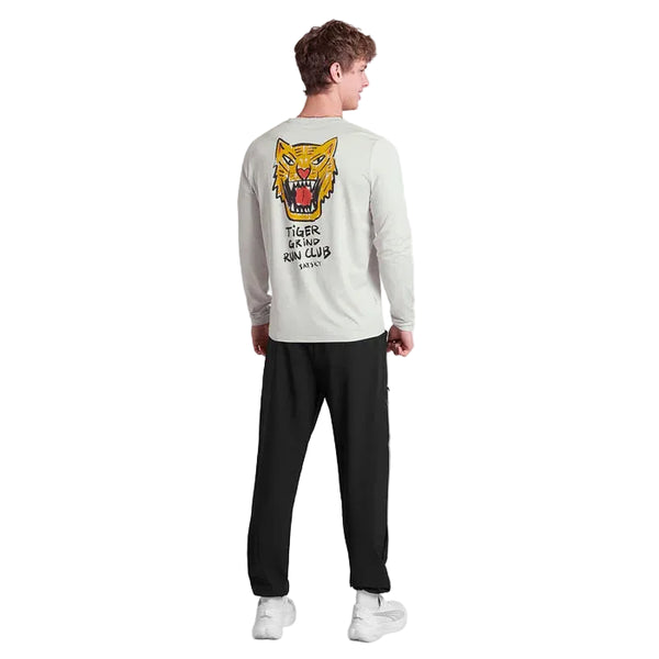 En mand står og kigger væk, iført en grå Saysky Tiger Pace Long Sleeve (herre) med et stort illustreret tigerhoved og teksten "TIGER GLAND RUN CLUB" på ryggen, sammen med sorte bukser og hvide sneakers.