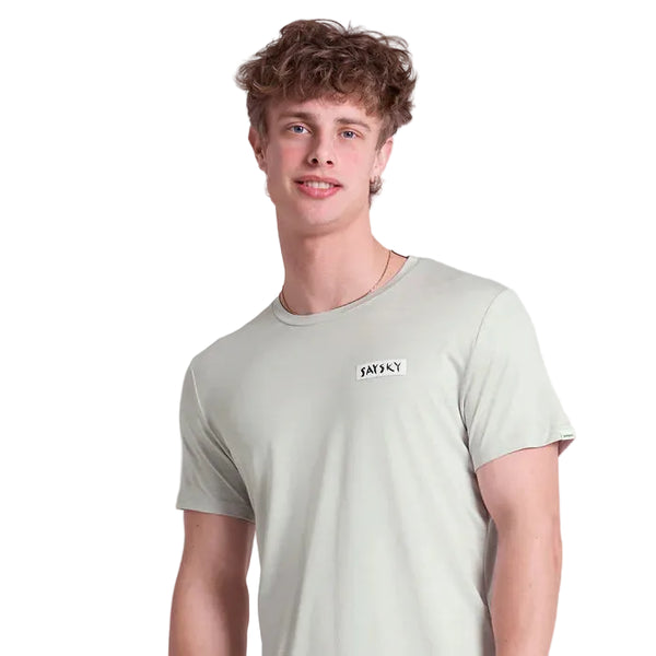 En ung mand med krøllet brunt hår bærer Saysky Tiger Pace T-shirt, Herre, Grey - et hurtigtørrende træningstrøje - mens han står på en almindelig hvid baggrund og ser lige frem med et neutralt udtryk.