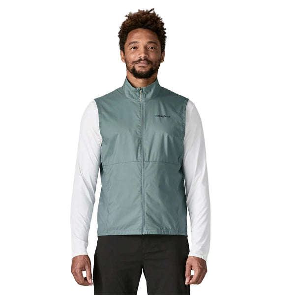 En mand iført en Patagonia Trail Craft Vest, Herre, i Blue Sage over en langærmet hvid skjorte og sorte bukser står med front mod en almindelig hvid baggrund.