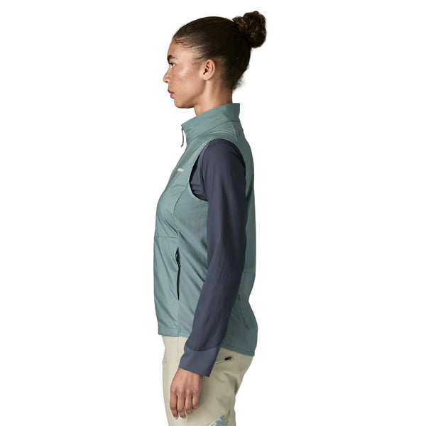 En kvinde i profil ser til venstre og er iført Patagonia Trail Craft Vest, Dame, Blue Sage over en mørk langærmet skjorte og lyse bukser. Vesten har en høj krave og lommer med lynlås. Hendes hår er bundet tilbage i en knold.