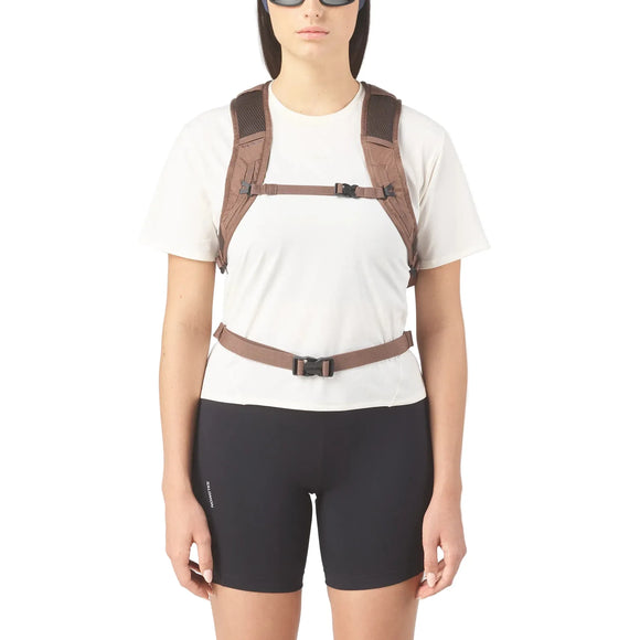 En person i hvid t-shirt og sorte shorts iført Salomon Trailbazer 10 unisex-rygsæk i Mustang/Black Coffee med to brystremme og en taljerem står foran en almindelig hvid baggrund.