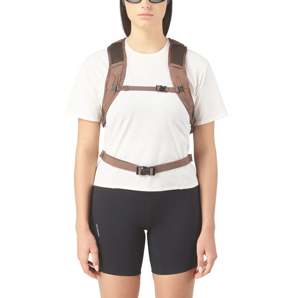 En person i hvid t-shirt og sorte shorts iført Salomon Trailbazer 10 unisex-rygsæk i Mustang/Black Coffee med to brystremme og en taljerem står foran en almindelig hvid baggrund.