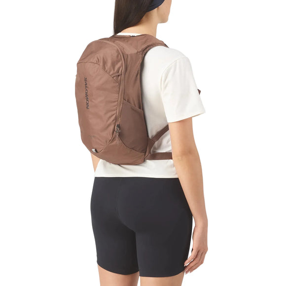 En person i hvid t-shirt, sorte shorts og en brun Salomon Trailbazer 10 (Unisex, Mustang/Black Coffee)-rygsæk står med ansigtet vendt væk fra kameraet.