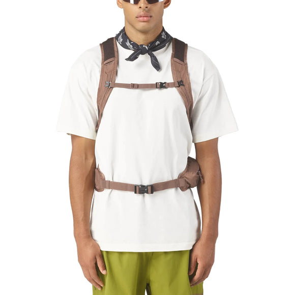 En person i hvid t-shirt, grønne bukser, sort halstørklæde, solbriller og brunt seletilbehør - ideel til vandreture med Salomon Trailbazer 20 Unisex-rygsækken i Mustang/Black Coffee.