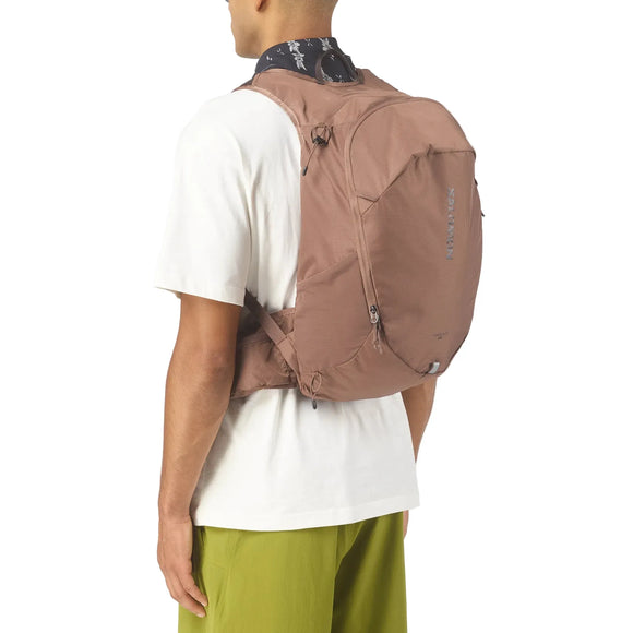 En person iført en Salomon Trailbazer 20 unisex rygsæk i Mustang/Black Coffee over en hvid t-shirt og grønne shorts, set bagfra og fra siden mod en ensfarvet baggrund - klar til vandretur.