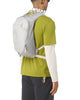 En person iført en grøn T-shirt med korte ærmer over en hvid langærmet skjorte, mørke bukser og en Salomon Trailblazer 10 Unisex-rygsæk i farverne Cloudburst/Paloma/Silver, fotograferet bagfra foran en ensfarvet baggrund.