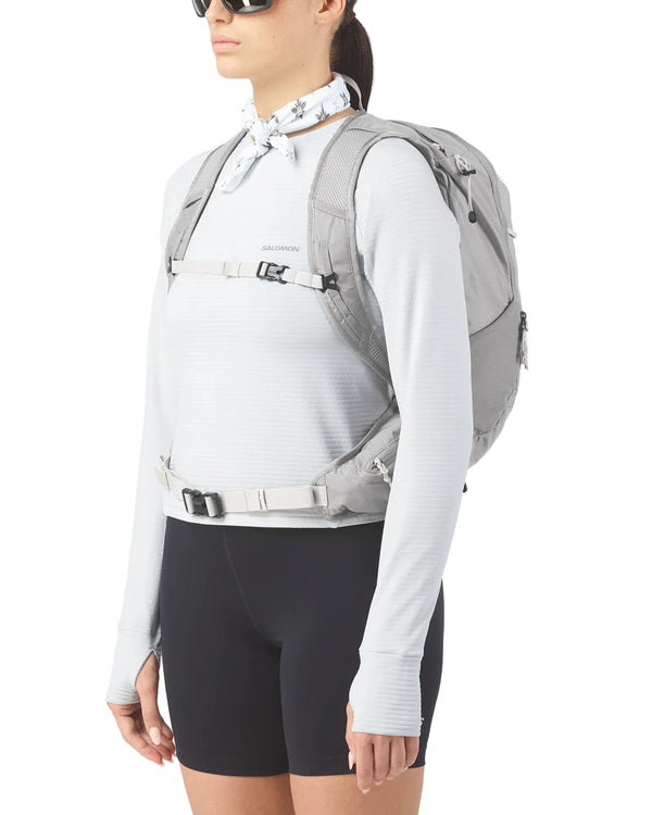 Der ses en person med solbriller, en hvid langærmet trøje, sorte shorts og et hvidt bandana, der har Salomon Trailblazer 20 (Unisex, Cloudburst/Paloma/Silver) på, stående foran en ensfarvet hvid baggrund.
