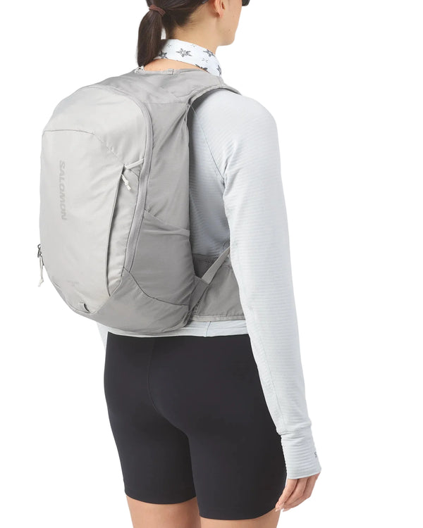 En person set bagfra bærer en Salomon Trailblazer 20-rygsæk (Cloudburst/Paloma/Silver), der er perfekt til løbependling, sammen med en hvid langærmet trøje, sorte shorts og en mønstret halsvarm – alt sammen på en hvid baggrund.