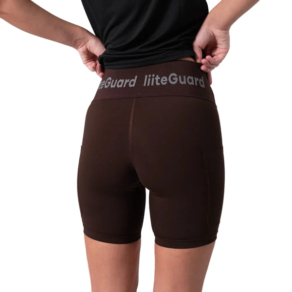 En person iført en sort T-shirt og Liiteguards »Training Tights Short, Dame, Coffee Bean« – hvor Liiteguard-logoet er synligt på linningen – står med ryggen til og retter på linningen med hænderne, hvilket fremhæver den perfekte pasform, der er typisk for højtaljede tights designet til kvinders træning.