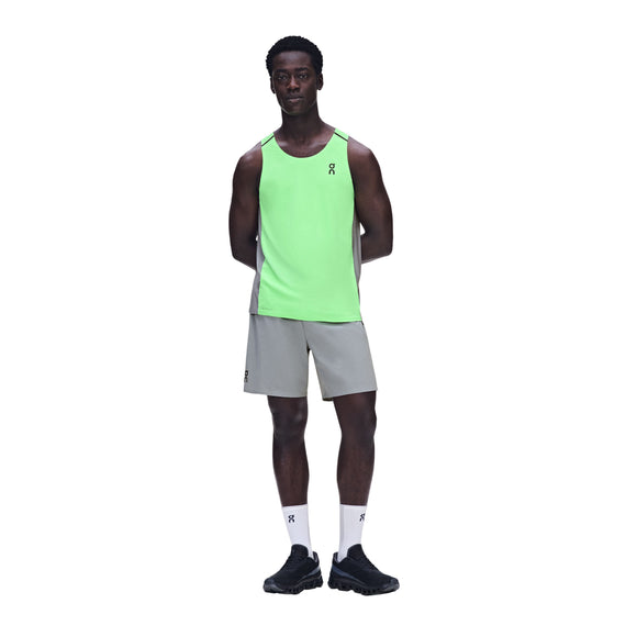 En person står med ansigtet fremad og armene bag ryggen, iført ON Performance Tank til mænd i farvekombinationen Verdite/Apollo, kombineret med lysegrå shorts, hvide sokker og sorte sportssko, foran en ensfarvet hvid baggrund.