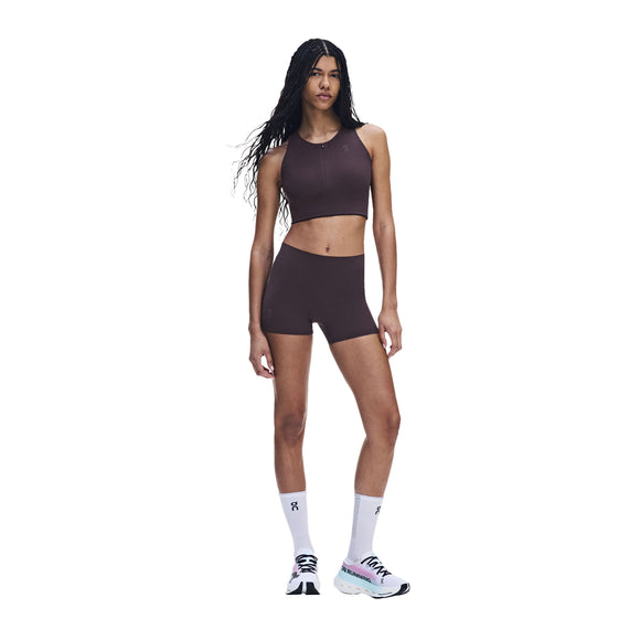 En person står foran en hvid baggrund iført ONs Zero Short Tights til kvinder i farven Urchin, kombineret med en matchende mørkebrun kort sportstop. Personen har langt flettet hår, hvide ankelstrømper og tykke hvide sneakers.
