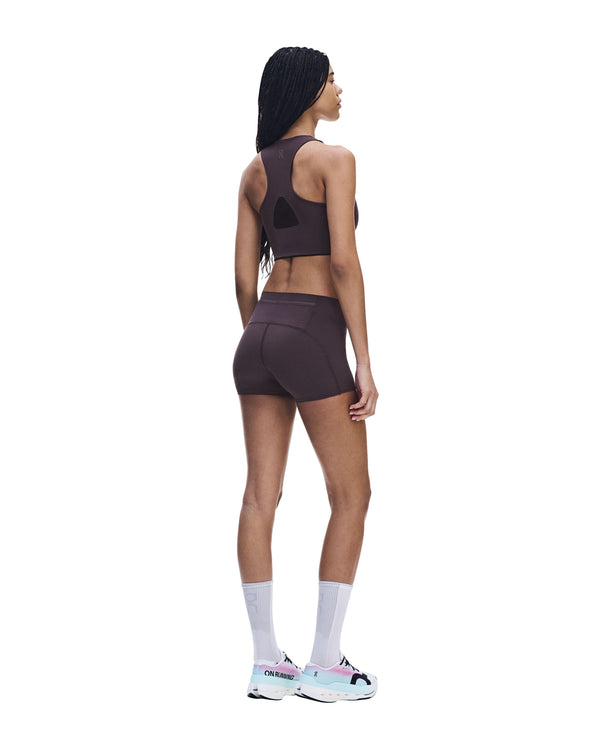 En kvinde står med ryggen til kameraet iført ON Zero Short Tights til kvinder i farven Urchin, kombineret med en matchende sports-BH, hvide sokker og hvide sportssko med detaljer i lyserødt og blågrønt, foran en hvid baggrund.