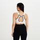 En kvinde med langt mørkt hår er iført en Shock Absorber Ultimate Run Bra Padded, Dame, White - en svedtransporterende sports-bh til maksimal støtte - og sorte leggings, vist bagfra mod en almindelig hvid baggrund.