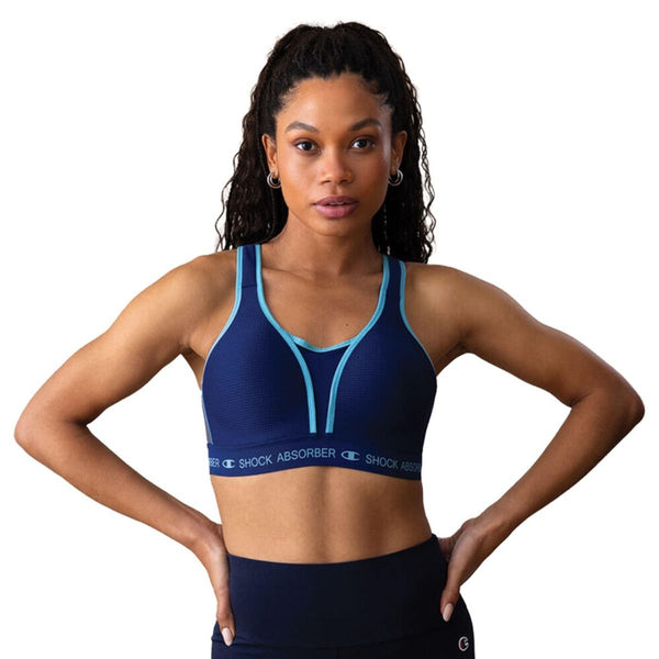 En kvinde med langt krøllet hår bærer Shock Absorber Ultimate Run Bra Padded, Dame, i Sail Away Navy, mens hun står med hænderne på hofterne mod en hvid baggrund.