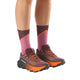 En person iført Salomon Ultra Glide 4 W trail-løbesko til kvinder (Huckle/Black/Redor) og lilla, læglange strømper med et let geometrisk mønster, der er perfekte til lange distancer, står på en hvid baggrund.