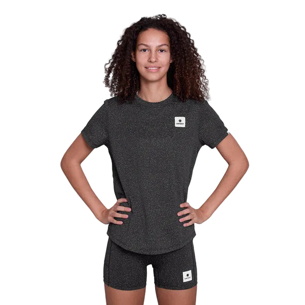 En kvinde står med ansigtet fremad og hænderne på hofterne, iført Saysky Universe Combat T-Shirt, Dame, Black Universe og matchende shorts med små rektangulære logoplapper. Dette bliver hurtigt en favorit for dem, der søger hurtigtørrende løbetøj. Baggrunden er almindelig hvid.