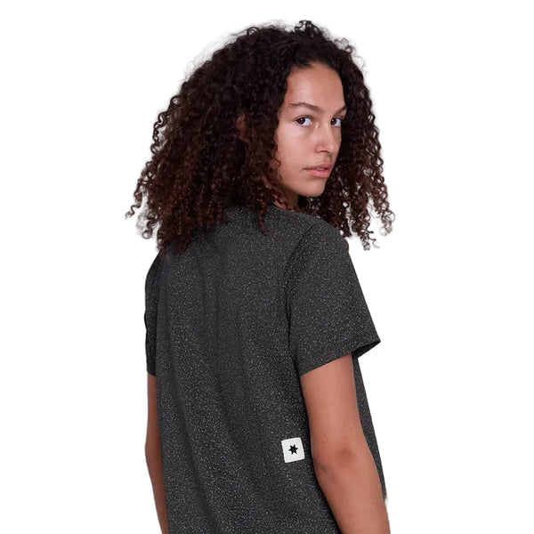 En kvinde med krøllet brunt hår bærer Saysky Universe Combat T-Shirt, Dame, Black Universe - en kortærmet, hurtigtørrende løbetøj med en lille hvid plet på ryggen - og står på en almindelig hvid baggrund og kigger over skulderen på kameraet.