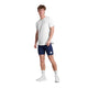 En mand kigger fremad mod en hvid baggrund iført Saysky Universe Combat T-Shirt, Herre, White Universe, sammen med marineblå sportsshorts, hvide sokker og hvide sneakers. Hans udtryk er neutralt.