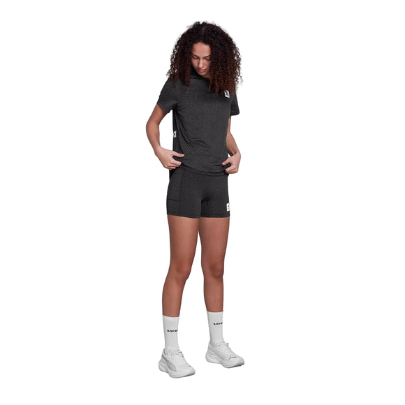 En person med krøllet hår er iført Saysky Universe Combat+ Short Tights 4", Dame, Black Universe, en matchende mørk t-shirt, hvide sokker og hvide sneakers, mens han står på en hvid baggrund og kigger ned med hænderne nær linningen.