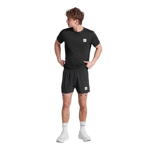 En ung mand står på en hvid baggrund iført Saysky Universe Pace Shorts 5" i Black Universe, en sort t-shirt, hvide sokker og hvide sneakers. Han kigger til venstre med hænderne på hofterne.