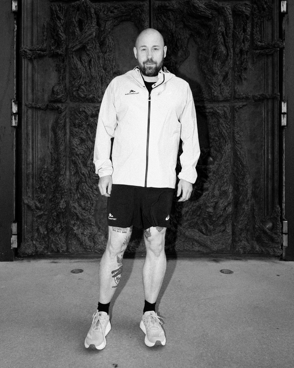 En skaldet mand med skæg er iført en lys jakke med lynlås, sorte shorts og sportssko. Han står foran en stor, struktureret dør. Hans ben har synlige tatoveringer. Billedet er i sort-hvid.