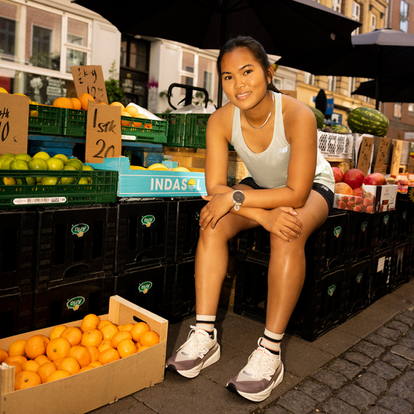 En kvinde i sportstøj sidder på en kasse ved en udendørs frugtbod, omgivet af kasser med appelsiner, lime og andre produkter. Hun ser på kameraet med et afslappet udtryk. Markedet ligger på en gade i byen.