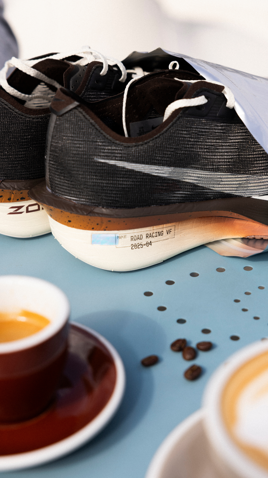 Et nærbillede af et par sorte løbesko med hvide såler placeret på en blå flade ved siden af en rød kaffekop og underkop. Kaffebønner og kaffedråber er spredt på bordet.