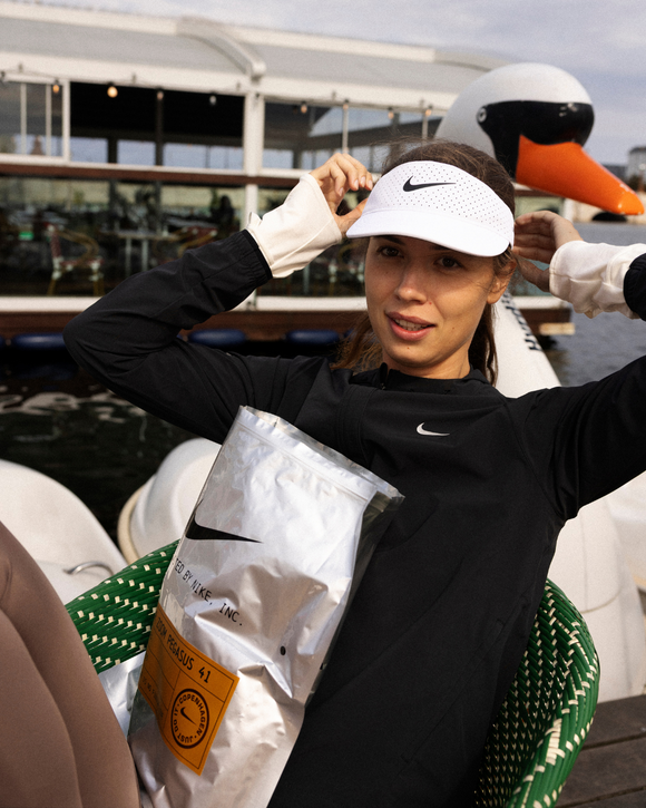 En kvinde i sort Nike-jakke og hvidt Nike-visir justerer sit visir, mens hun sidder udendørs på en grøn stol. Hun holder en sølvfarvet taske med tekst, og i baggrunden ses en stor svaneformet padlebåd.
