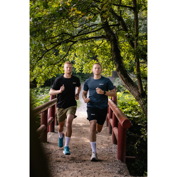 To mænd jogger side om side på en grussti over en rød træbro, omgivet af grønne træer. Begge er iført Liiteguard Ground-Tech T-shirt, Herre, Black og løbesko, som sikrer komfort og ydeevne under deres træning.