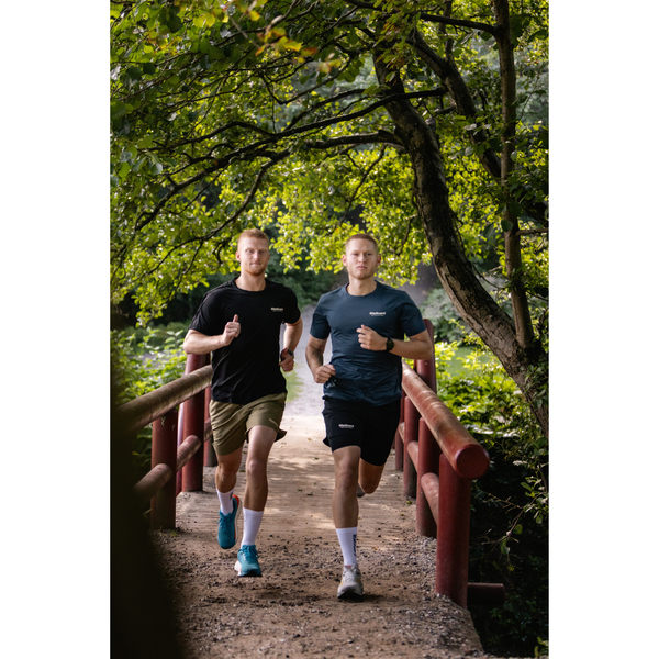 To mænd løber side om side på en grøn sti over en træbro, begge iført LiiteGuard Ground-Tech T-shirt, Herre, Black-svedtransporterende og ideel til herre træning i det fri.