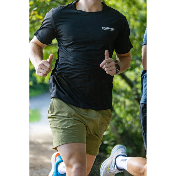 En person jogger på en sti i LiiteGuards Ground-Tech T-shirt, Herre, Black og olivenfarvede shorts. Den svedtransporterende tee holder dem komfortable midt i det grønne, hvor kun deres overkrop og ben er synlige.